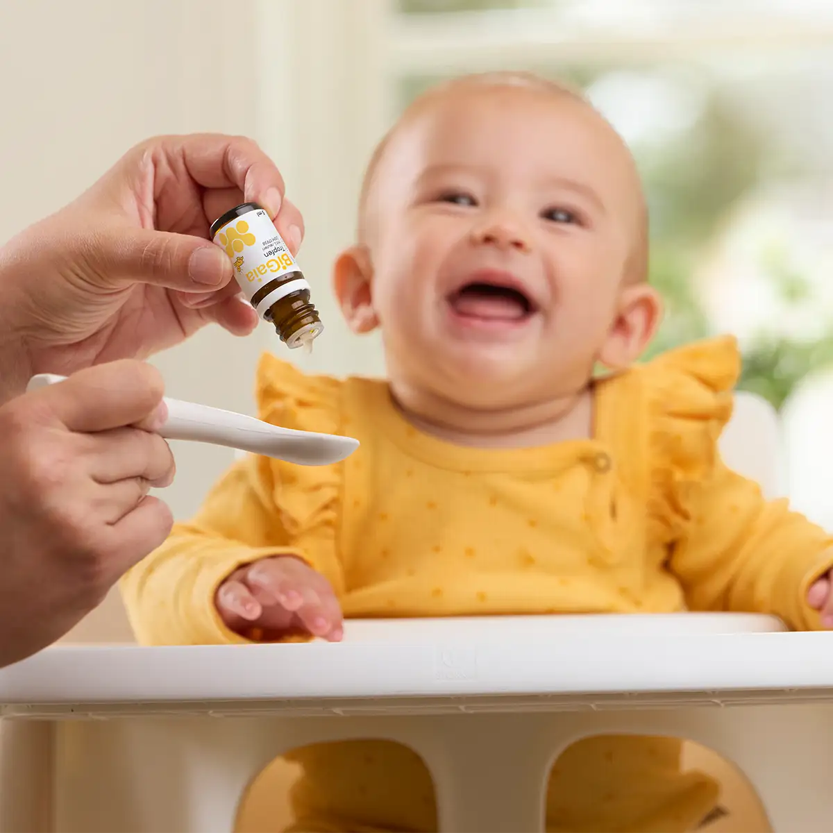 Ein lachendes Baby im Hochstuhl bekommt BiGaia Tropfen per Löffel gereicht.